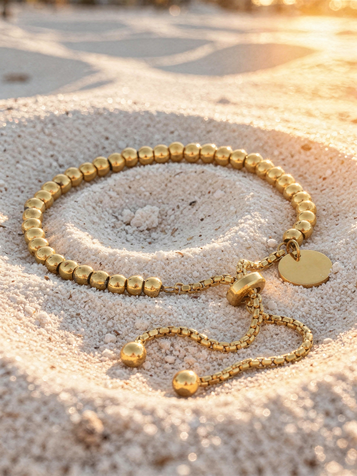 Perla Beaded Bracelet in 18k gold-plated stainless steel resting on fine white beach sand at golden hour