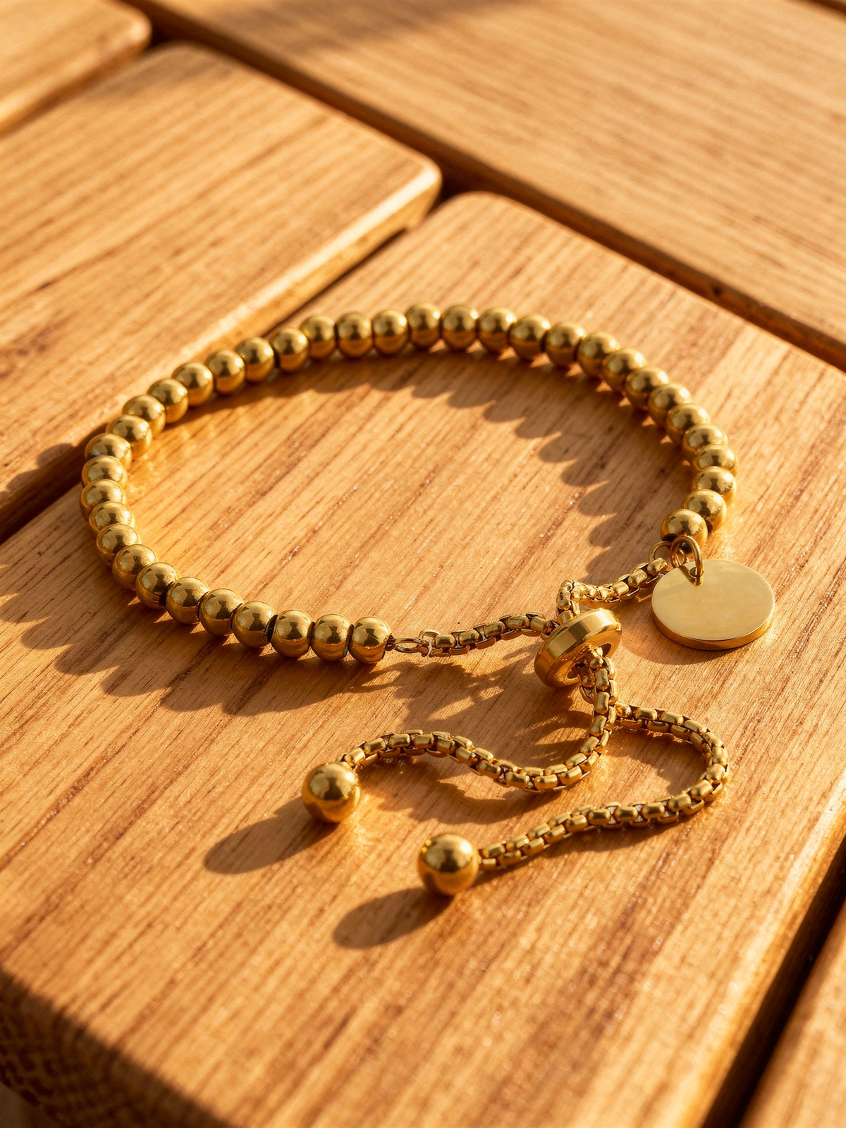 Perla Beaded Bracelet in 18k gold-plated stainless steel resting on a pale teak wood deck at golden hour